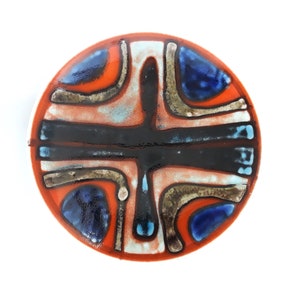 May include: A round ceramic tile with a geometric design in shades of orange, blue, brown, and black. The design features a cross shape with curved lines and a white background.