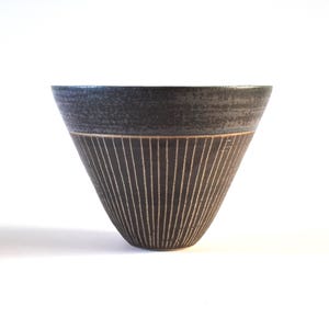 May include: A dark grey ceramic bowl with a textured rim and vertical etched lines. The bowl has a conical shape and a contrasting band of lighter colour near the top. The background is white.