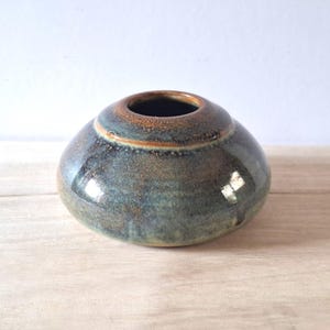 May include: A small, round ceramic vase with a wide opening. The vase has a glossy finish with shades of blue, green, and brown. The vase is on a wooden surface.