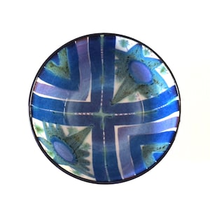 May include: A round, decorative bowl with a dark rim. The bowl's interior features a blue and white geometric design with intersecting lines and floral-like shapes. The color palette includes shades of blue and green.