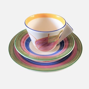 May include: A colourful teacup and saucer set with a whimsical design. The cup has a yellow rim and a pink and green floral pattern. The saucer has a multi-coloured striped design.