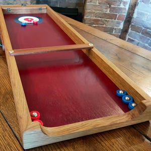 May include: A wooden tabletop game with a red playing surface and a target in the shape of a bullseye. The game includes red and blue playing pieces.