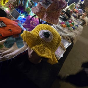 May include: A yellow crocheted bucket hat with a large white eye and green pupil. The hat is on a mannequin head.
