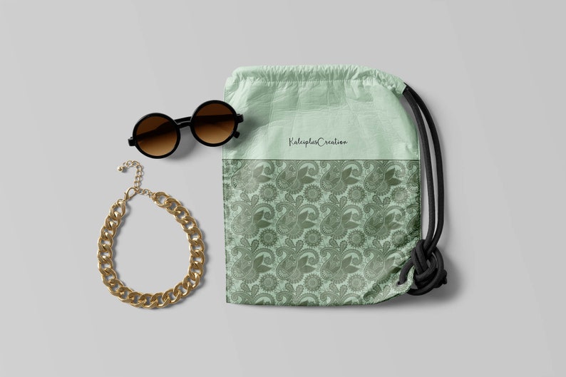 May include: A green drawstring bag with a paisley pattern and the text "Kaleidoscope Creation". The bag is next to a pair of round sunglasses and a gold chain necklace.