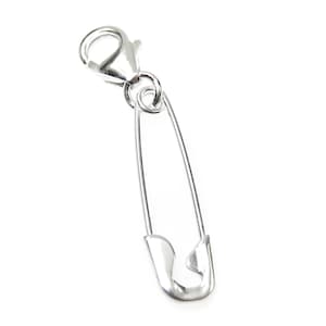 May include: A silver-colored safety pin charm with a lobster clasp. The safety pin is a classic design, with a curved pin and a protective clasp. The charm is made of a shiny, reflective metal and is set against a white background.
