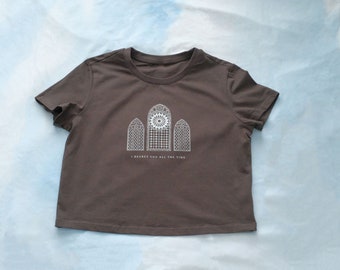 Puede incluir: Una camiseta marrón recortada con un gráfico blanco de tres ventanas de vidrieras. El texto "I regret you all the time" está impreso debajo del gráfico.