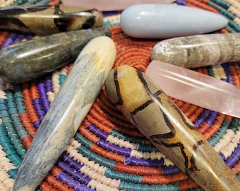 May include: A collection of polished stone wands in various colors, including pink, blue, and brown, arranged in a woven basket with a colorful pattern. The wands have smooth, rounded shapes and varying patterns.