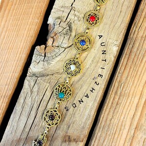 May include: Gold-toned bracelet with intricate circular designs, each holding a different colored gemstone: turquoise, red, blue, white, teal, and dark red. The bracelet includes a Hamsa hand charm. The text "AUNTIE 2 HANDS" is visible.