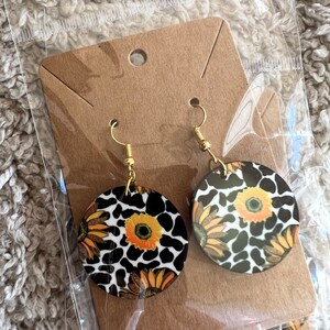 May include: A pair of wooden earrings with a black and white cow print design and yellow sunflowers. The earrings are on a brown card with a hole in the top for hanging.