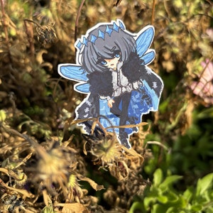 Peut inclure: Un autocollant représentant un personnage de style anime avec des ailes bleues, une couronne et une cape sombre. Le personnage a les cheveux gris et se détache sur un fond de fleurs séchées et de feuillage vert. L'autocollant a une bordure blanche.