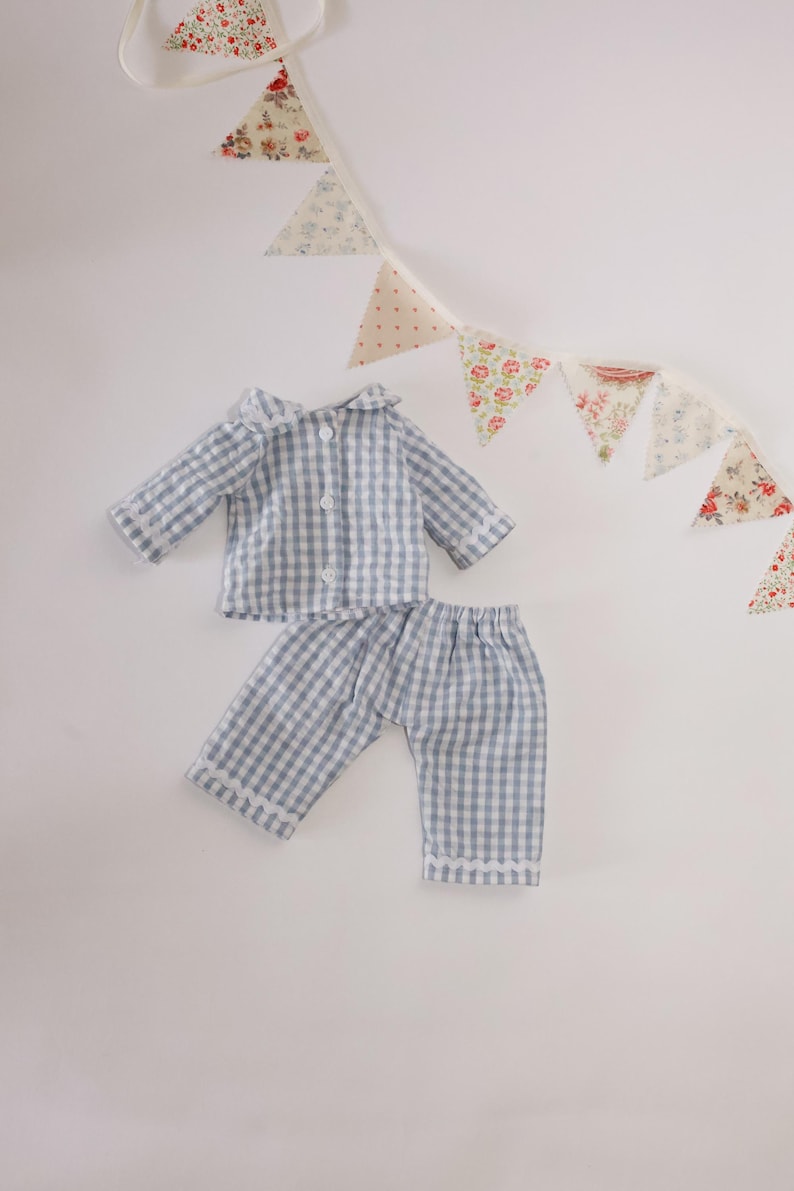 May include: A blue and white checked pyjama set, comprising a collared shirt and trousers. The set is displayed on a white background, with a decorative garland of floral and polka dot fabric triangles.