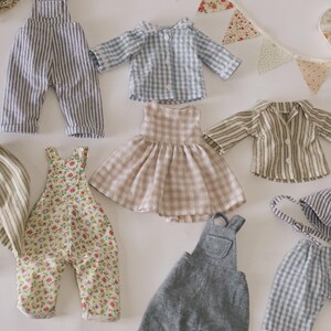 May include: A selection of miniature doll clothes in various styles and patterns. The garments include striped dungarees, a gingham shirt, a floral print dress, and a striped shirt. A matching hat and a decorative banner are also present.