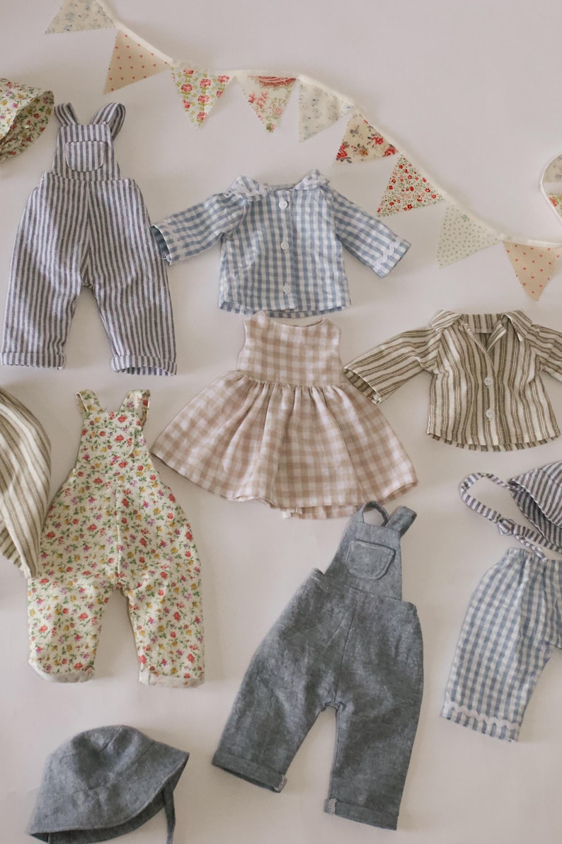 May include: A selection of miniature doll clothes in various styles and patterns. The garments include striped dungarees, a gingham shirt, a floral print dress, and a striped shirt. A matching hat and a decorative banner are also present.