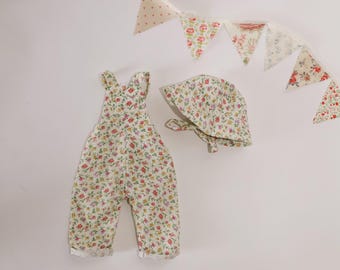 May include: A baby's floral print outfit set, comprising dungarees and a bonnet, with a matching floral banner. The outfit has a white base with a colourful floral pattern. The banner features triangle flags with floral and polka dot patterns.