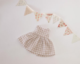 May include: A beige and white gingham dress with a flared skirt is positioned below a garland of floral and polka dot fabric triangles. The garland is suspended from a white ribbon.