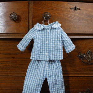 May include: A blue and white gingham pajama set, comprising a button-down top and matching trousers, hangs on a hook. The pajamas have white lace trim and a white collar. The set is displayed against a wooden chest of drawers.