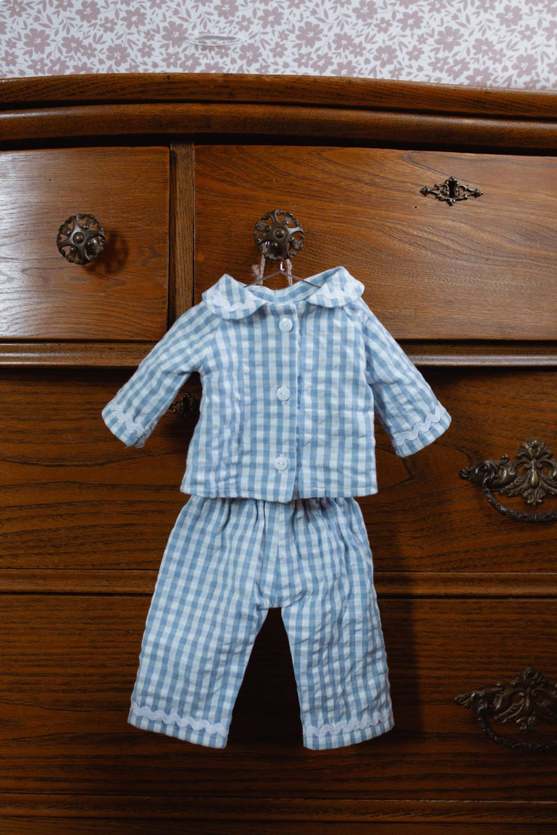 May include: A blue and white gingham pajama set, comprising a button-down top and matching trousers, hangs on a hook. The pajamas have white lace trim and a white collar. The set is displayed against a wooden chest of drawers.