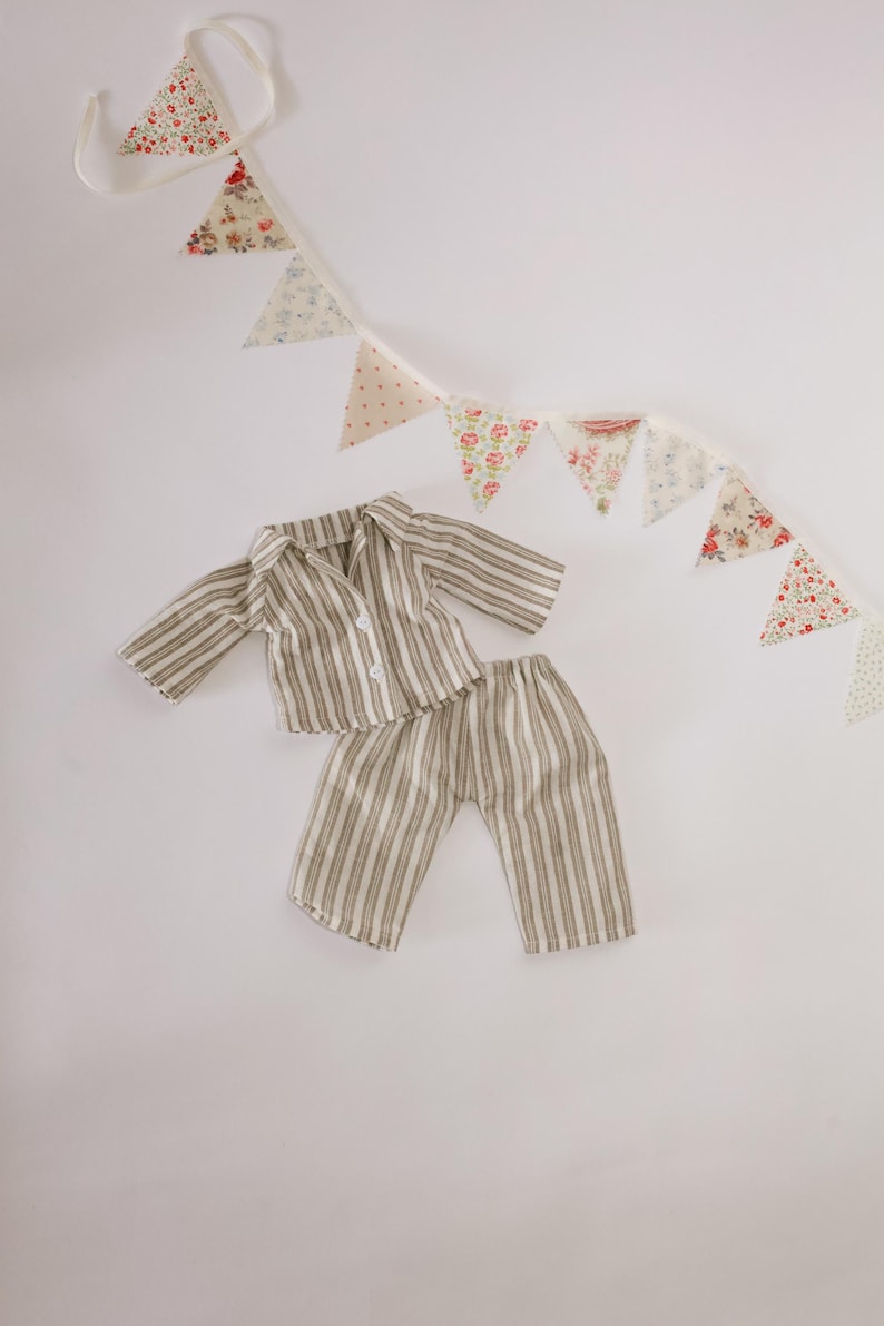 Pode incluir: Um conjunto de pijama em miniatura com um padr&atilde;o listrado em tons de bege e castanho. O conjunto inclui uma camisa com bot&otilde;es e cal&ccedil;as a condizer. Uma grinalda de tri&acirc;ngulos de tecido com estampados florais e de bolinhas est&aacute; pendurada por cima.