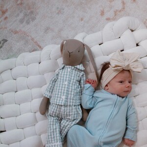 May include: A baby in a light blue ribbed onesie and a cream bow headband lies beside a stuffed bunny in blue and white checked pyjamas. The items are on a white woven surface, with a patterned rug in the background.