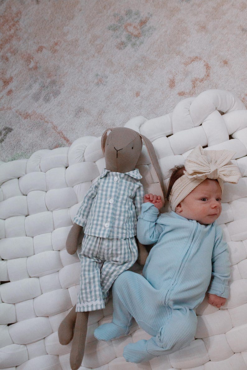 May include: A baby in a light blue ribbed onesie and a cream bow headband lies beside a stuffed bunny in blue and white checked pyjamas. The items are on a white woven surface, with a patterned rug in the background.