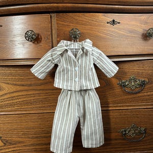 Pode incluir: Um conjunto de pijama em miniatura com um padr&atilde;o listrado em bege, branco e castanho. O conjunto inclui uma camisa com bot&otilde;es e cal&ccedil;&otilde;es a condizer, expostos num cabide. O pijama &eacute; projetado para uma boneca ou figura pequena.
