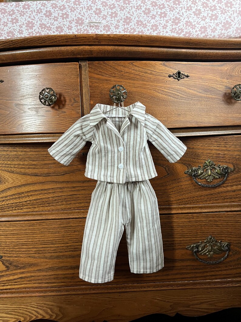 Pode incluir: Um conjunto de pijama em miniatura com um padr&atilde;o listrado em bege, branco e castanho. O conjunto inclui uma camisa com bot&otilde;es e cal&ccedil;&otilde;es a condizer, expostos num cabide. O pijama &eacute; projetado para uma boneca ou figura pequena.