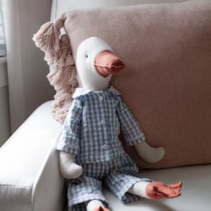 May include: A soft goose toy dressed in blue and white gingham pyjamas with a white collar. The goose has a white head, a peach-coloured beak and feet, and is seated on a white chair next to a pink cushion.
