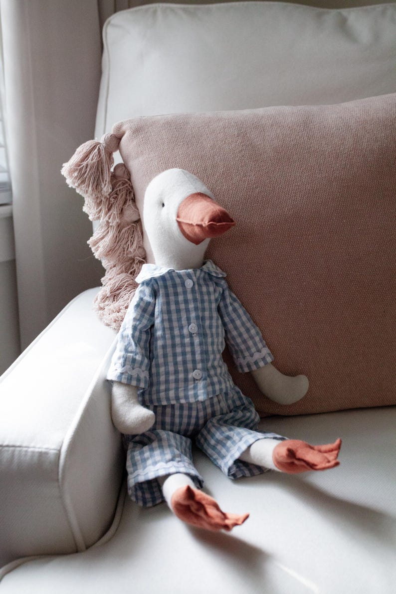 May include: A soft goose toy dressed in blue and white gingham pyjamas with a white collar. The goose has a white head, a peach-coloured beak and feet, and is seated on a white chair next to a pink cushion.