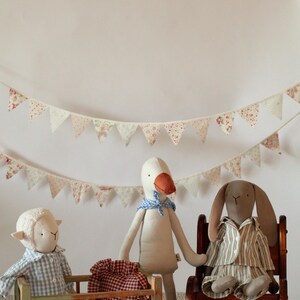 May include: A nursery scene featuring three soft toys: a sheep in a cot, a goose, and a rabbit in a rocking chair. A red and white checked bag is near the goose. A garland of floral pennants hangs above.