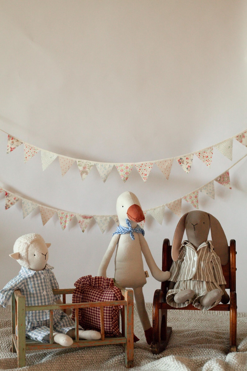 May include: A nursery scene featuring three soft toys: a sheep in a cot, a goose, and a rabbit in a rocking chair. A red and white checked bag is near the goose. A garland of floral pennants hangs above.