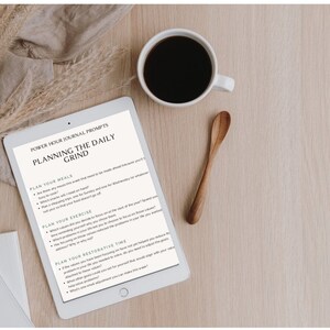 May include: A white tablet with a black screen displaying a journal prompt titled "Planning the Daily Grind". The prompt includes sections for planning meals, exercise, and restorative time. The tablet is on a wooden surface with a cup of coffee and a wooden spoon.
