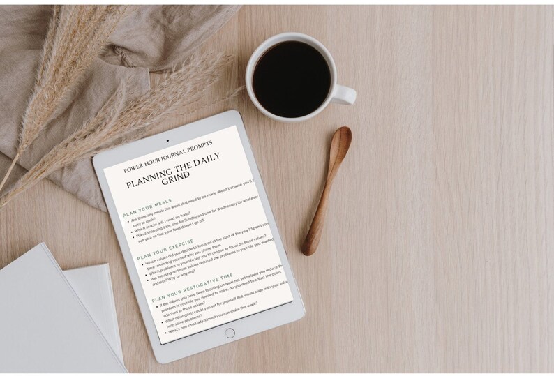 May include: A white tablet with a black screen displaying a journal prompt titled "Planning the Daily Grind". The prompt includes sections for planning meals, exercise, and restorative time. The tablet is on a wooden surface with a cup of coffee and a wooden spoon.