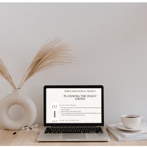 May include: A laptop computer with a white background showing a screen with the text "POWER HOUR JOURNAL PROMPTS PLANNING THE DAILY GRIND" and two numbered bullet points: "01 PLAN YOUR MEALS" and "02 PLAN YOUR EXERCISE". The laptop is on a wooden table with a vase of dried pampas grass, a pair of glasses, and a white coffee cup and saucer.