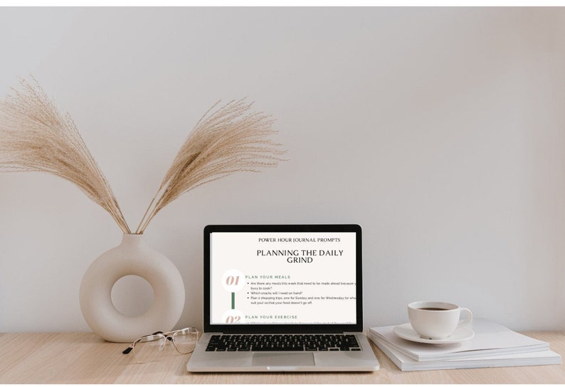 May include: A laptop computer with a white background showing a screen with the text "POWER HOUR JOURNAL PROMPTS PLANNING THE DAILY GRIND" and two numbered bullet points: "01 PLAN YOUR MEALS" and "02 PLAN YOUR EXERCISE". The laptop is on a wooden table with a vase of dried pampas grass, a pair of glasses, and a white coffee cup and saucer.