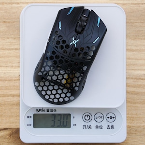 May include: A black computer mouse with a honeycomb shell design and blue accents resting on a white digital scale. The scale display shows a weight of 33.0 grams. The mouse features a scroll wheel and a central logo on its top surface.