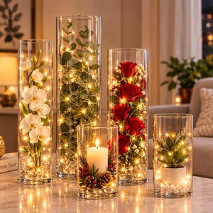 May include: Five clear cylindrical glass vases containing decorative arrangements. One holds white orchids, one green eucalyptus, one red roses, one a white pillar candle with pinecones, and one a small potted succulent, all illuminated by string lights.
