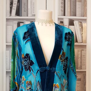 May include: A teal blue velvet and sheer chiffon jacket with a floral pattern. The jacket has a V-neckline and a button closure. The jacket is on a white mannequin.