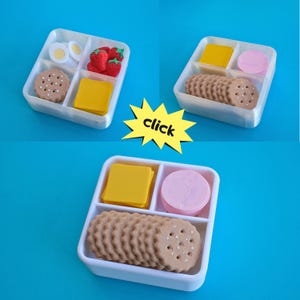 May include: A white plastic lunchbox with compartments, filled with miniature food items. Includes yellow cheese squares, pink circles, brown cookies, and red strawberries. The image also contains the word "click" in a yellow starburst.