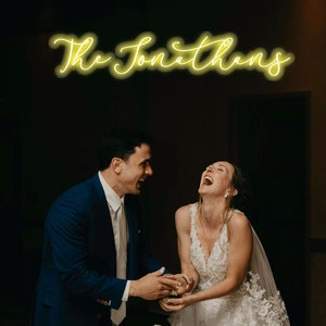 May include: A bride and groom laughing as they cut a white wedding cake with a single white rose on top. The cake is on a white tablecloth. The words "The Jonathans" are written in yellow neon script above the couple.