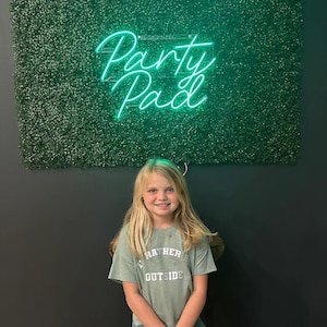 Könnte beinhalten: Ein junges Mädchen mit langen blonden Haaren steht vor einer grünen Wand mit einem Neon-Schild, auf dem "Party Pad" steht. Sie trägt ein grünes T-Shirt mit dem Aufdruck "I'd Rather Be Outside".