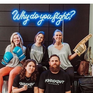 May include: A group of people wearing boxing gloves and shirts with the words "Knockout Fitness" and "Bumble" on them. The group is standing in front of a black wall with a neon sign that says "Why do you fight?"