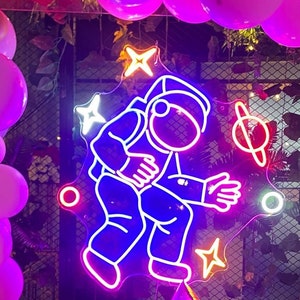 Könnte beinhalten: Ein Neon-Schild mit einem Astronauten in einem Raumanzug, umgeben von einem Planeten und Sternen. Das Schild hängt an einer Decke, die mit rosa und goldenen Ballons geschmückt ist.