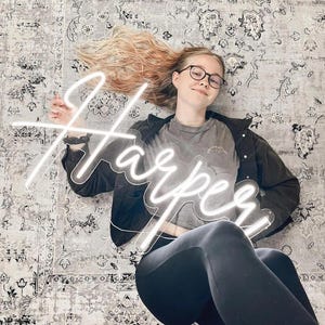 Puede incluir: Un letrero de ne&oacute;n blanco que muestra el nombre "Harper" en una fuente cursiva. El letrero lo sostiene una persona que usa gafas, una camiseta gris y leggings negros. El fondo es una alfombra estampada.