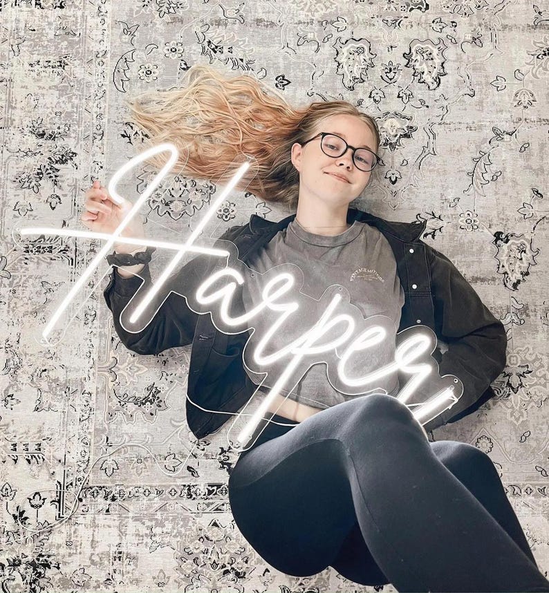Puede incluir: Un letrero de ne&oacute;n blanco que muestra el nombre "Harper" en una fuente cursiva. El letrero lo sostiene una persona que usa gafas, una camiseta gris y leggings negros. El fondo es una alfombra estampada.
