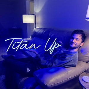 May include: A person sitting on a couch with a blue neon sign that says "Titan Up".