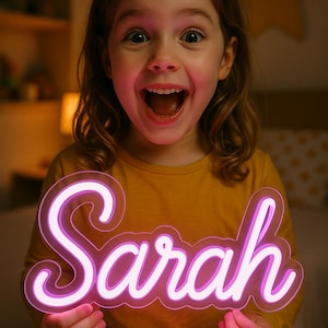 Puede incluir: Una ni&ntilde;a sostiene un letrero de ne&oacute;n con el nombre "Sarah" en una fuente cursiva. El letrero brilla con una luz rosa y blanca. La ni&ntilde;a lleva una camiseta de manga larga de color amarillo mostaza y tiene una amplia sonrisa emocionada.
