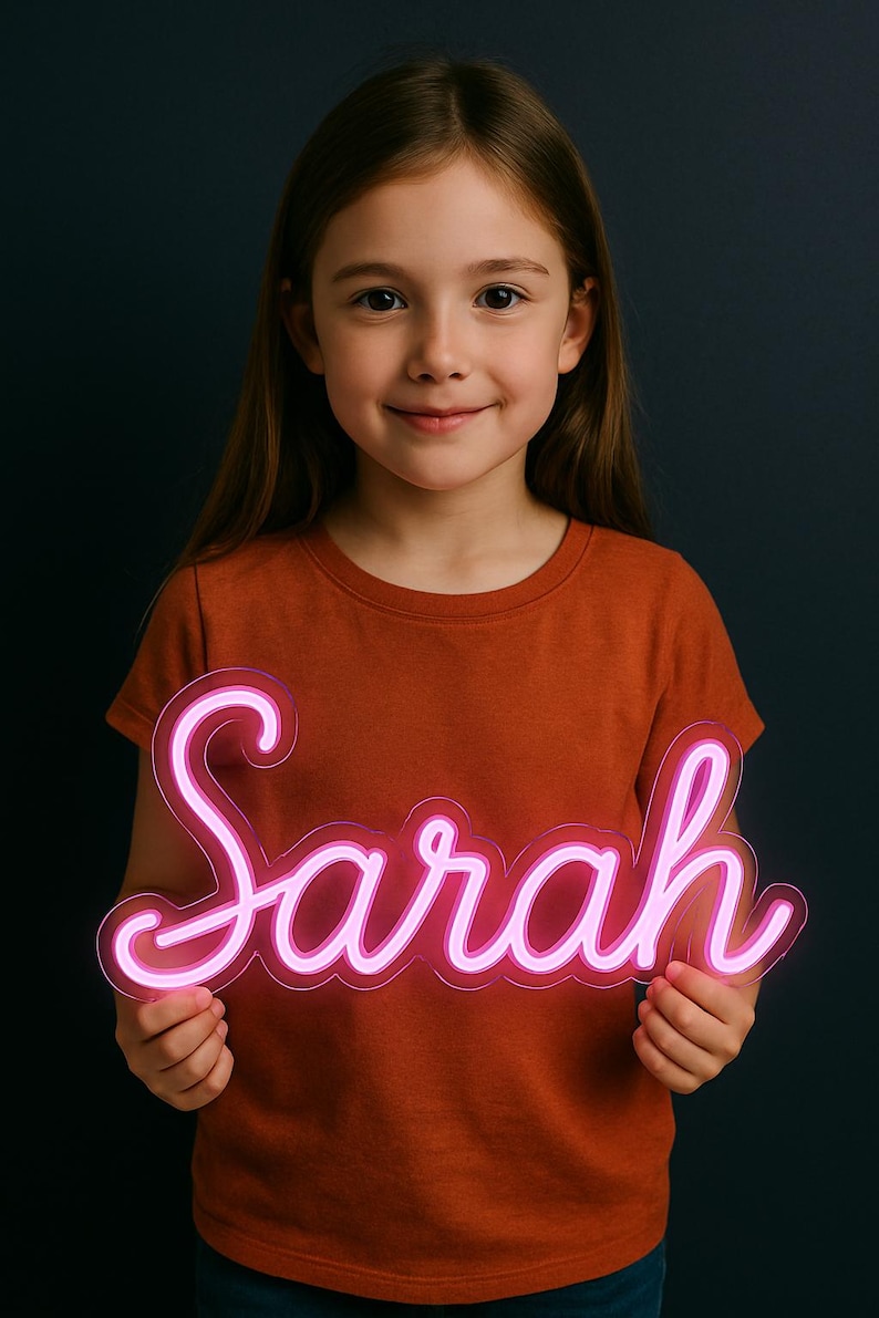 Puede incluir: Un letrero de ne&oacute;n rosa con el nombre "Sarah". El letrero lo sostiene una persona que lleva una camiseta naranja. El fondo es azul oscuro.