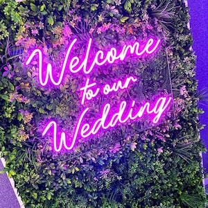 Può includere: Un'insegna al neon con la scritta "Welcome to our Wedding" in un carattere corsivo, illuminata in rosa acceso. L'insegna è posta su uno sfondo di lussureggiante fogliame verde con tocchi di fiori viola, creando un'esposizione vibrante e accogliente.
