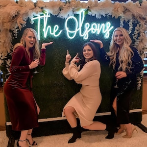 以下が含まれることがあります： 3人の女性が、「The Olsons」と書かれたネオンサインのある緑色の壁の前でポーズをとっています。真ん中の女性はベージュのセーターとスカートを着用しており、両側の女性はドレスを着用しています。左側の女性はワインレッドのベルベットのドレスを着用しており、右側の女性は黒いスパンコールのドレスを着用しています。