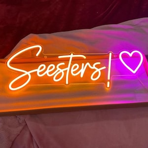 May include: Neon sign with the word "Seesters!" in white script with an orange glow and a pink heart with a white outline.